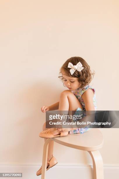 menina de 3 anos vestida com um vestido pastel shift e arco branco, sentado em uma cadeira colocando seus sapatos - sapato bege - fotografias e filmes do acervo