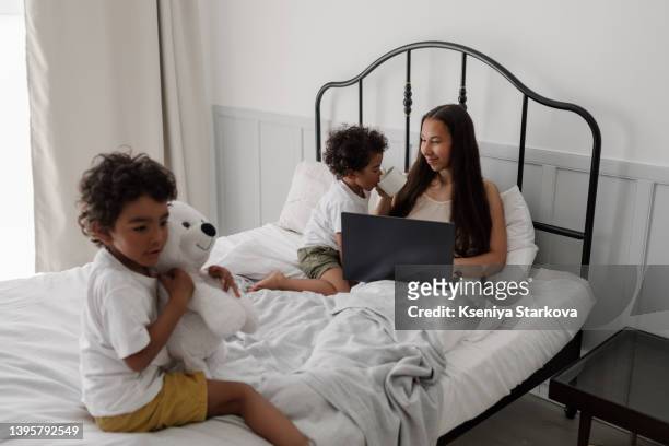 young beautiful asian woman with long dark hair sits in bed working on a laptop next to her two little dark-skinned sons who play next to each other on the bed, morning awakening - 3 4 length business woman ストックフォトと画像