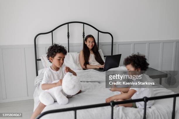 young beautiful asian woman with long dark hair sits in bed working on a laptop next to her two little dark-skinned sons who play next to each other on the bed, morning awakening - 3 4 length business woman stock pictures, royalty-free photos & images