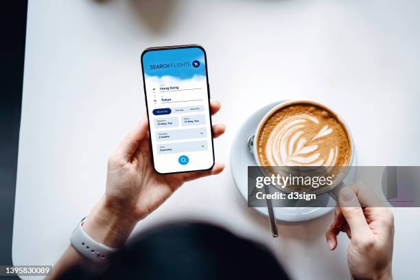 overhead view of young woman sitting on desk booking flight tickets on airline website online with smartphone while having a cup of coffee. travel planning. booking a holiday online. business person planning business trip - life search stock pictures, royalty-free photos & images