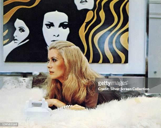Catherine Deneuve, French actress, laying on a fur rug, on which sits a telephone, with an artwork hanging on the wall behind her, in a publicity...