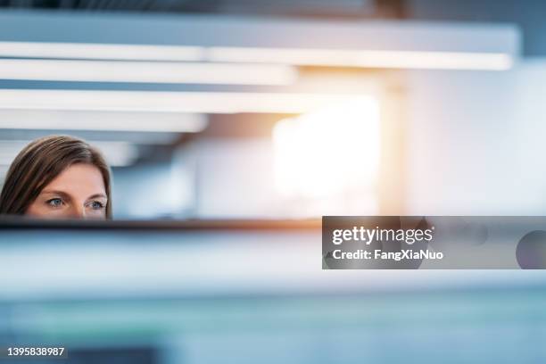 woman looking across office from behind cubicle wall and computer monitor - kantoorpolitiek stockfoto's en -beelden