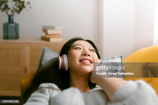 front view of a relaxed and smiling asian woman enjoying listening to music with wireless headphones while lying on a sofa at home. - funeral parlor fotografías e imágenes de stock