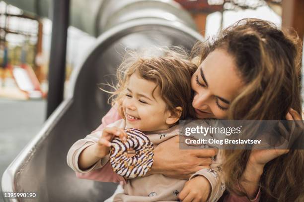 mother and daughter laughing and kissing on playground. harmony in family. baby holding sweet donut - kinderspielplatz stock-fotos und bilder