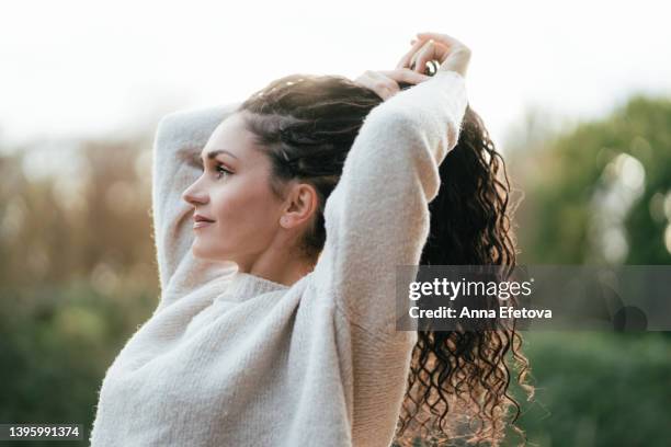 beautiful authentic woman is fixing her long curly hair. she is wearing cozy knitted sweater. concept of natural beauty - candid beautiful young woman face stock pictures, royalty-free photos & images