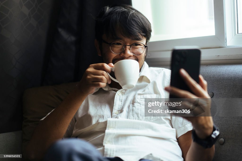 Close up positive emotion Asian young adults enjoy and relax by drinking coffee and using their phones on the sofa at home Domestic life.