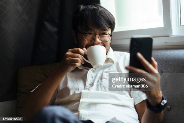 close up positive emotion asian young adults enjoy and relax by drinking coffee and using their phones on the sofa at home domestic life. - asian journalist stock pictures, royalty-free photos & images