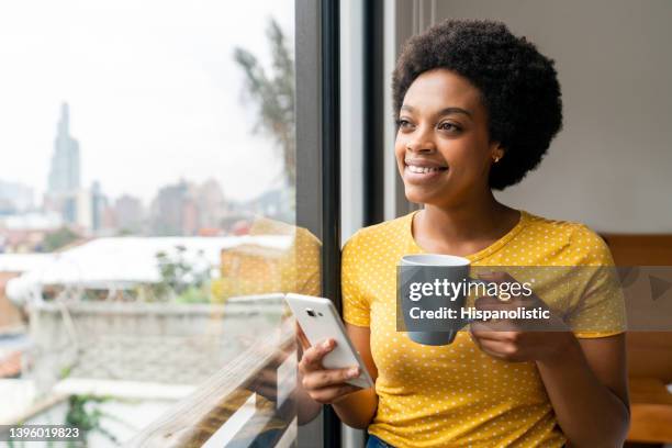 mujer feliz en casa tomando café mientras usa su teléfono - arrendatario fotografías e imágenes de stock