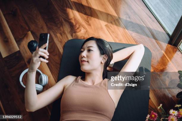 high angle shot of fitness young asian sports woman relaxing after working out, lying on yoga mat and using her smartphone against sunlight in the morning, reusable water bottle and headphones by her side. health, fitness and wellness concept - asian exercise stock-fotos und bilder