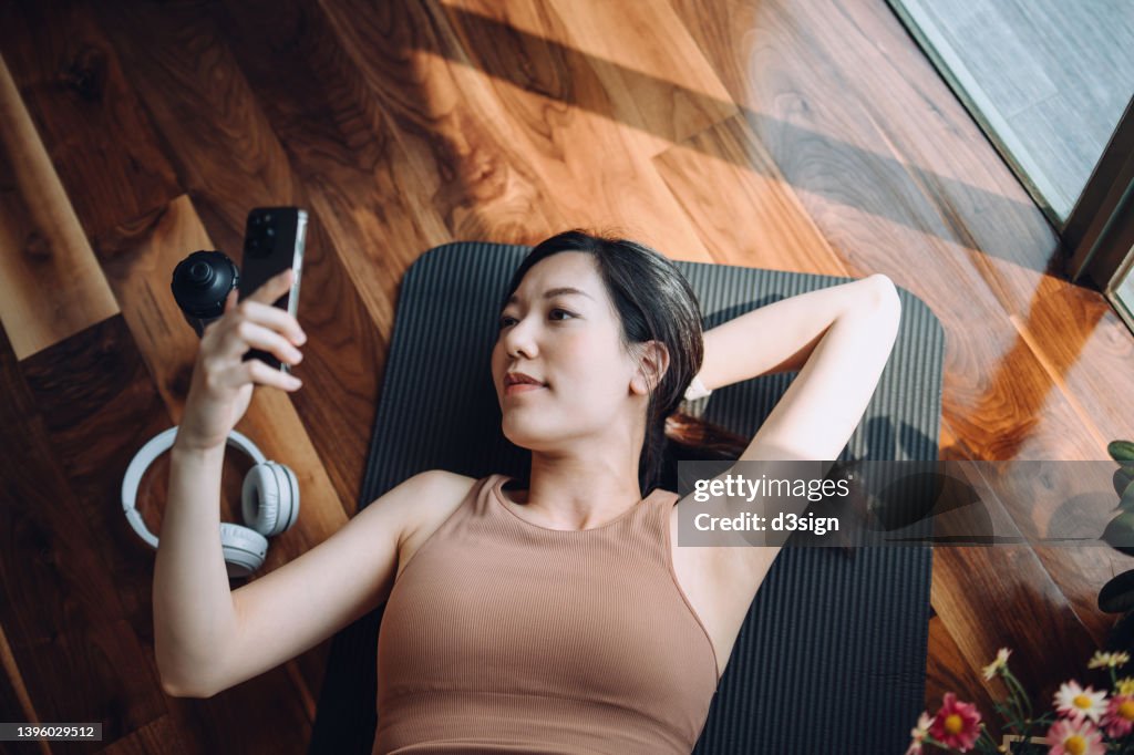 High angle shot of fitness young Asian sports woman relaxing after working out, lying on yoga mat and using her smartphone against sunlight in the morning, reusable water bottle and headphones by her side. Health, fitness and wellness concept