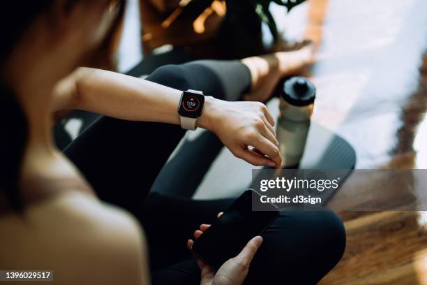 over the shoulder view of fitness asian woman checking pulse and training performance on fitness tracker on smartwatch after exercising at home. recording health status with smartphone. health and fitness training with technology. healthy living lifestyle - fitness tracker stock-fotos und bilder
