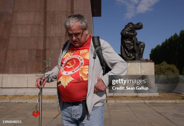 Man wearing a t-shirt that shows a communist hammer and sickle and reads, in Russian: "Patriotic War" walks with a carnation past a sculpture of a...