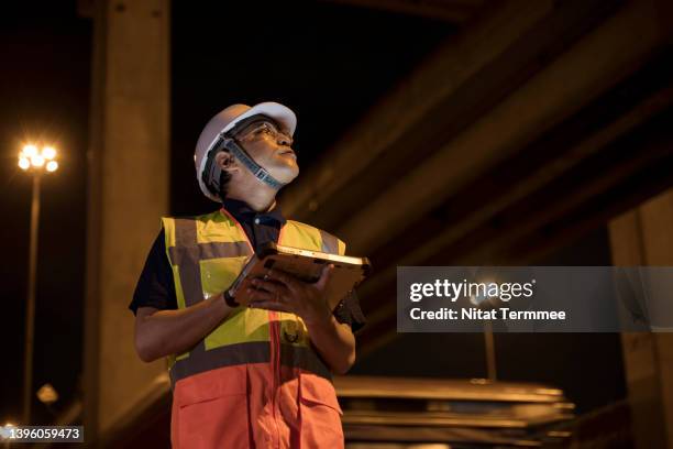 heavy civil and transportation construction services. low angle view of a japanese project manager looking onto crossroad or highway during survey for construction project while holding a tablet at night shift. - civil engineer - fotografias e filmes do acervo