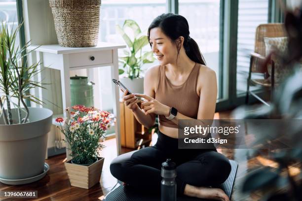 confident fitness young asian woman relaxing after exercising at home. young asian female in sports wear scrolling on smartphone and resting after home work out. fitness training with technology. health, fitness and wellness concept - asian and indian ethnicities smartwatch phone stock pictures, royalty-free photos & images