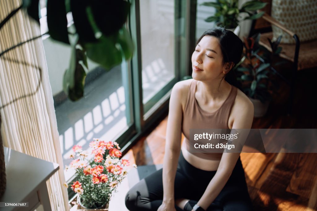 Active young Asian sports woman taking a break after working out at home, sitting on exercise mat taking a deep breath with her eyes closed. Sports and exercise routine. Health, fitness and wellness concept