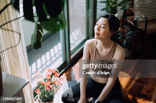 Active young Asian sports woman taking a break after working out at home, sitting on exercise mat taking a deep breath with her eyes closed. Sports and exercise routine. Health, fitness and wellness concept Active young Asian sports woman taking a break after working out at home, sitting on exercise mat taking a deep breath with her eyes closed. Sports and exercise routine. Health, fitness and wellness concept