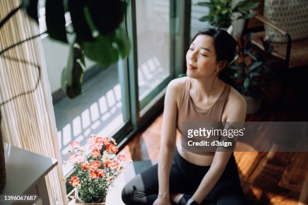 active young asian sports woman taking a break after working out at home, sitting on exercise mat taking a deep breath with her eyes closed. sports and exercise routine. health, fitness and wellness concept - salle yoga photos et images de collection