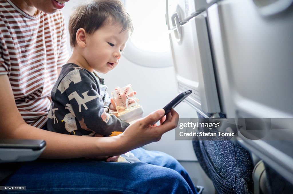 Mother taking airplane with a baby boy using cellphone for entertainment. Baby traveling in airplane flying sitting on his mother lap in the aircraft
