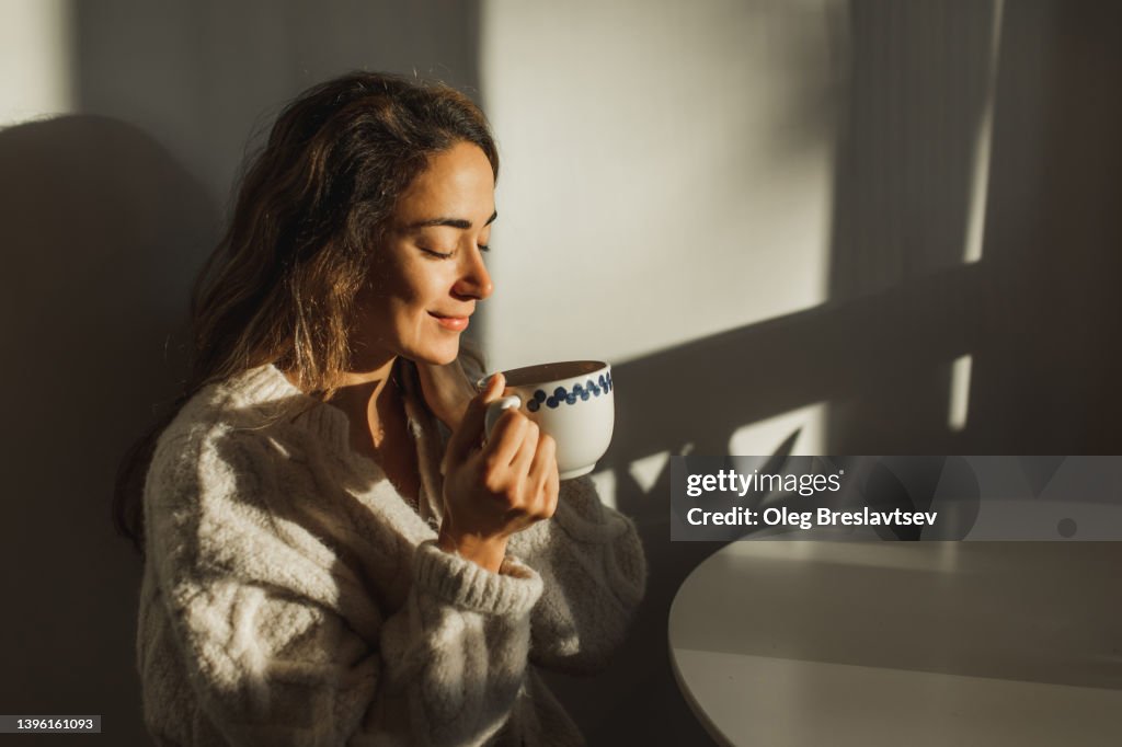 Young pretty woman enjoying morning coffee at home. Beautiful sun light in kitchen room.