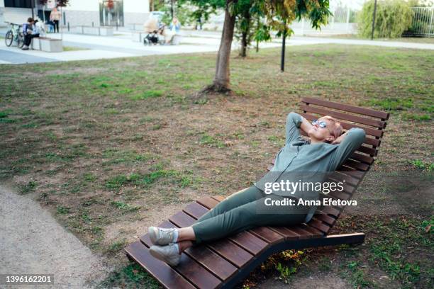 senior blonde woman in sunglasses is resting on wooden lounger in the city park with hands behind her head. she is wearing modern casual clothing - diva imagens e fotografias de stock