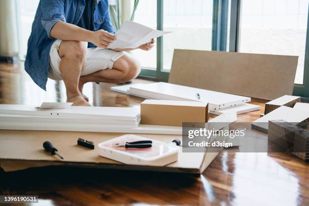 cropped shot of young asian man unboxing flat-pack furniture, reading instructions and preparing to assemble wooden furniture in his new apartment - manuale foto e immagini stock