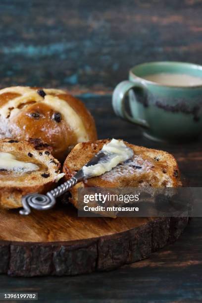 close-up image of two spiced buns on circular, wooden board, half a toasted, easter hot cross bun with melted butter and missing bite, butter knife, cup of tea, wood grain background, focus on foreground - raisin toast stock pictures, royalty-free photos & images