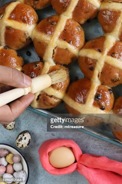 close-up image of freshly baked, homemade easter hot cross buns on greaseproof parchment paper lined baking tray, unrecognisable person using pastry brush to glaze buns with apricot jam, bowl of chocolate mini eggs, home baking concept, elevated view - hot cross bun stock pictures, royalty-free photos & images