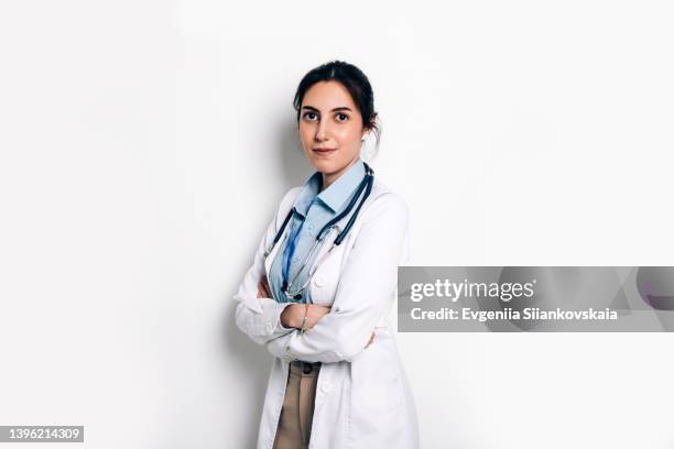 smiling woman doctor is wearing a lab coat and a stethoscope standing against white wall in medical office. - doctor lab coat stock-fotos und bilder