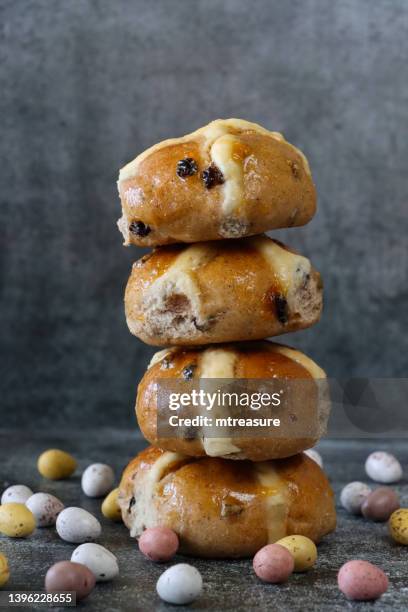 close-up image of stack of freshly baked, homemade easter hot cross buns surrounded by speckled candy coated chocolate mini easter eggs, mottled grey background, home baking concept, focus on foreground - hot cross bun stock pictures, royalty-free photos & images