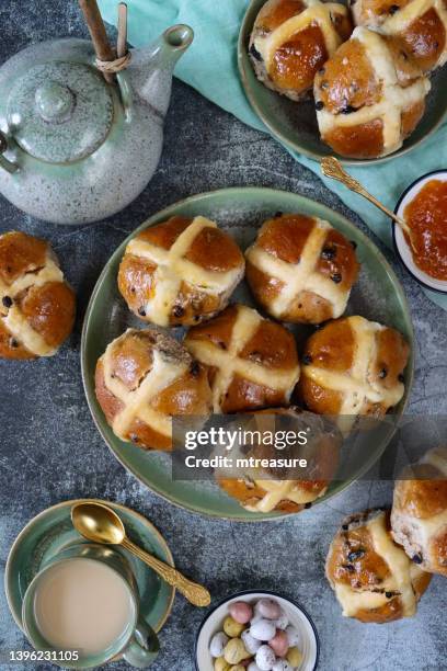 bild von hausgemachten oster-heißkreuzbrötchen auf tellern neben porzellan-teekanne und teetasse in untertasse mit teelöffel, gerichte aus aprikosenmarmelade und schokoladen-mini-eiern, grünem musselin, gesprenkeltem grauem hintergrund, erhöhte aussic - hot cross buns stock-fotos und bilder