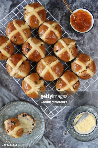 image of homemade easter hot cross buns on cooling rack, sliced and buttered spiced bun on plate, dish of apricot jam, butter dish and knife, mottled grey background, home baking concept, elevated view - hot cross bun stock pictures, royalty-free photos & images