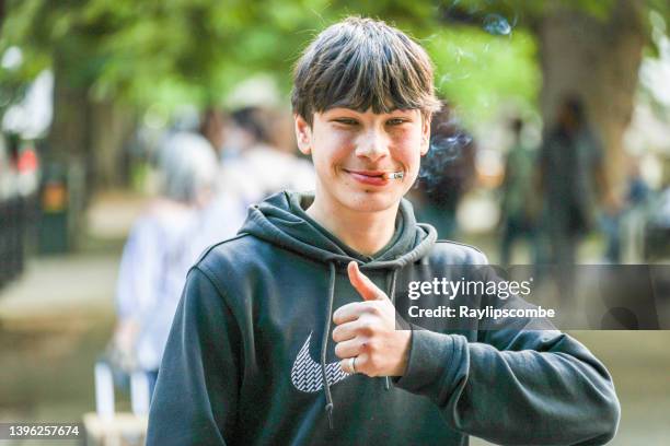 cheeky, grinning teenage boy posing for the camera with a thumbs up as a lit cigarette dangles from his mouth. - boys smoking cigarettes stock pictures, royalty-free photos & images