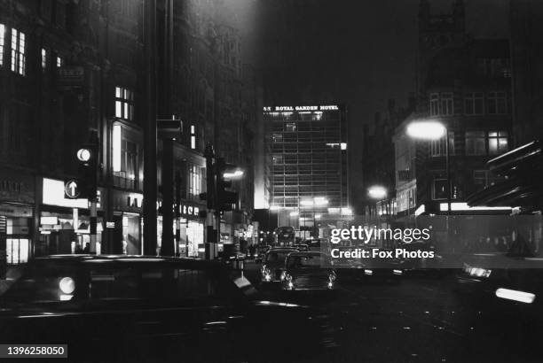 Illuminated shop fronts and traffic with the Royal Garden Hotel rising beyond on Kensington High Street, London, England, 1969. The hotel overlooks...
