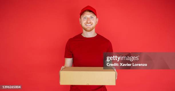 delivery man holding cardboard package over red isolated background - caja cartón fotografías e imágenes de stock