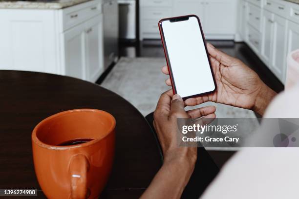 woman holds smart phone with blank screen while sitting at kitchen table - hands holding smart phone stock-fotos und bilder