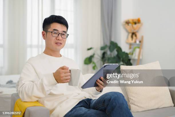 young man holding coffee cup and using tablet while sitting on sofa in home living room, looking at camera - social media profile stock pictures, royalty-free photos & images