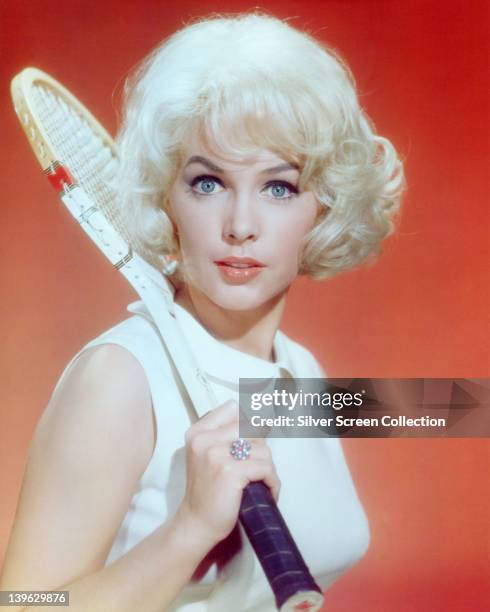 Stella Stevens, US actress, wearing a white sleeveless top and holding a wooden tennis racquet over her right shoulder in a studio portrait, against...