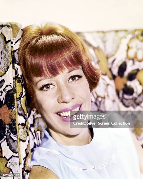 Headshot of Carol Burnett, US actress, singer and dancer, wearing pink lipstick, smiling while resting her head against a patterned cushion, circa...