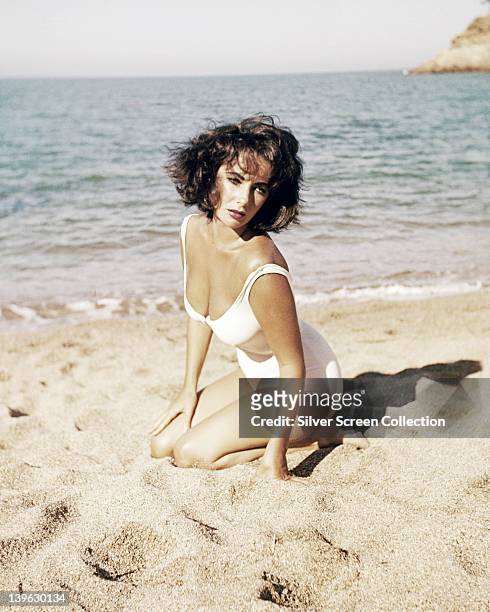 Elizabeth Taylor , British actress, wearing a white swimsuit as she kneels on the sands of a beach in a publicity still issued for the film,...
