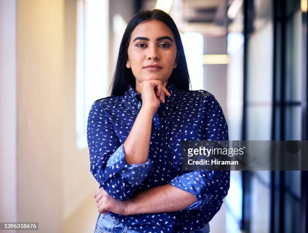 confident young indian businesswoman standing alone with her arms crossed and hand on her chin in the hallway of an office. one female only looking ambitious and determined at work - hand on chin stockfoto's en -beelden