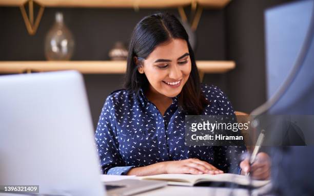 joven empresaria india escribiendo en un cuaderno mientras trabaja sola en una computadora portátil en una oficina. una mujer solo toma notas en su diario y planifica en línea mientras trabaja en su escritorio - arab businesswoman with books fotografías e imágenes de stock