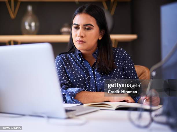 集中したインドのビジネスウーマンは、オフィスのラップトップで一人で仕事をしながら、ノートに書いています。ある女性は、自分の机で仕事をしながら、日記にメモをとり、オンライン� - confused writing ストックフォトと画像