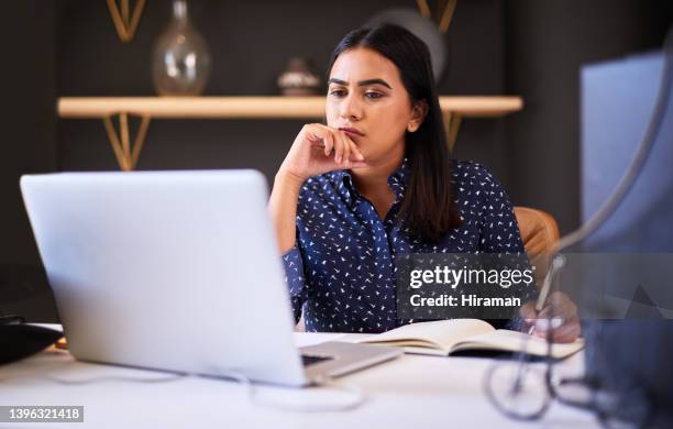 focused indian businesswoman writing in a notebook while working alone on a laptop in an office. one female only thinking before making notes in her diary and planning online while working at her desk - verward stockfoto's en -beelden