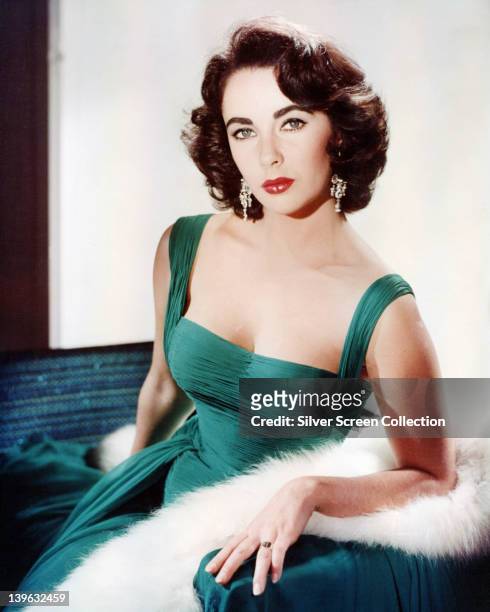 Elizabeth Taylor , British actress, wearing a green sleeveless low-cut dress, with a white fur wrap on the arm of the armchair in which she sits,...