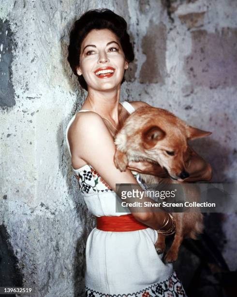 Ava Gardner , US actress, wearing a white dress with floral motifs, and a red belt, smiling as she holds a dog in her arms, in a studio portrait,...