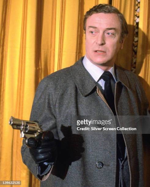 Michael Caine, British actor, wearing a grey overcoat and black leather gloves, pointing a handgun, as he stands before a yellow curtain in a...