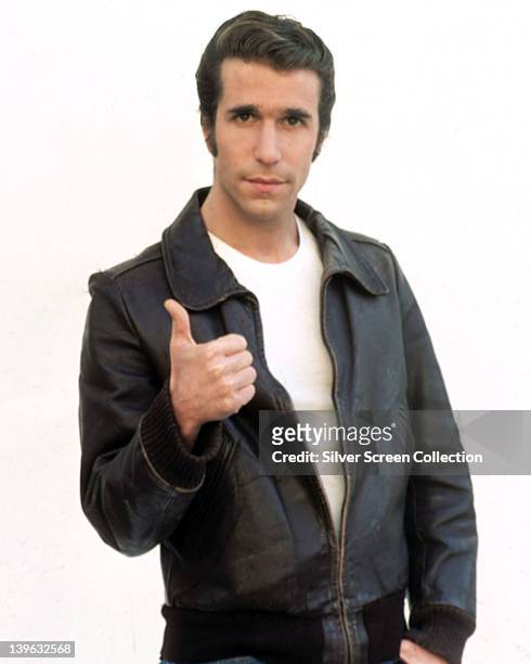 Henry Winkler, US actor, wearing a black leather jacket and white t-shirt, giving a thumbs up in a publicity portrait, against a white background,...