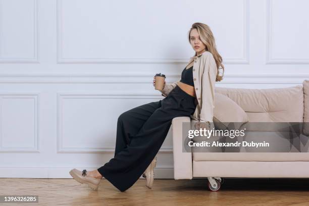 beautiful young blonde woman posing while sitting on the sofa.fashion model in stylish clothes in the studio looking at the camera. - beige fashion stock pictures, royalty-free photos & images