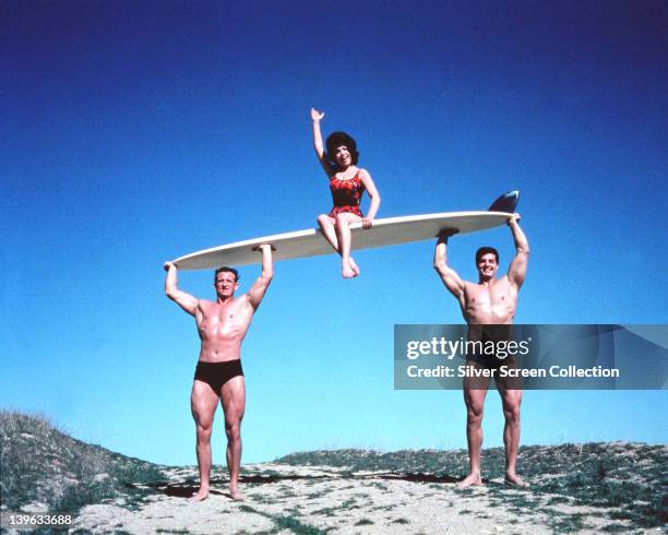 Annette Funicello, US actress and singer, wearing a red swimsuit, and waving while sitting on a surfboard which is being carried by two men in a...