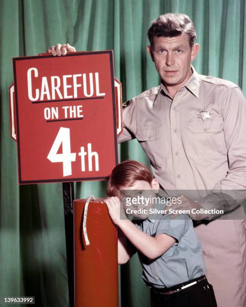 Andy Griffith, US actor, and Ron Howard, US actor, pose beside a large firework, with a sign reading 'Careful on the 4th' in a publicity portrait...